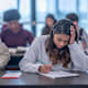 A female college student sits at her desk in a university classroom and takes notes. She has her head in her hand in concentration.