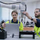 A Caucasian male and Caucasian female engineer are working on a robot in a lab. One of them is holding a laptop. The robot is blue and white.