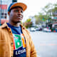 Black male HBCU college student wearing a cap and a blue sweatshirt with his school's HBCU logo walks downtown in a city on a fall afternoon.