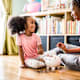 A Black mother and her young daughter are sitting on the floor in the living room at home. The mother is teaching her daughter about saving money as she helps count coins in her daughter's piggybank.