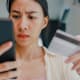 Asian American female college student using her credit card to pay her student loan bill. She is looking at her mobile phone, looking shocked at the amount she has to pay.