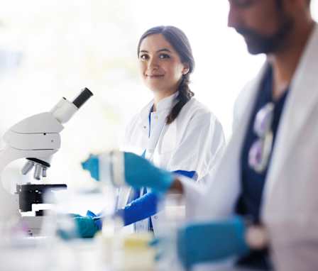 Two scientists working in a lab with a microscope wearing scrubs