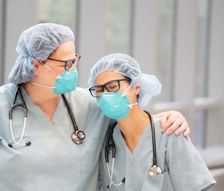 Two female, health care providers laugh while wearing light green medical scrubs, stethoscopes and N95 masks in an indoor hallway.