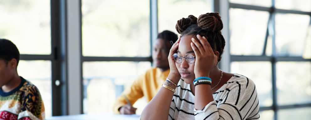 Worried young female student sitting with head in hands at desk during exam in community college classroom