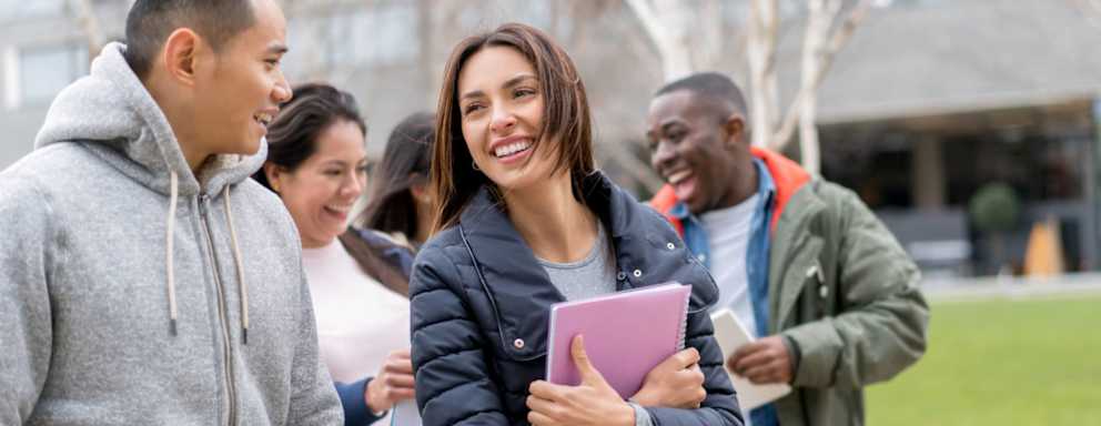 A small group of multi-ethnic international college students walking outside on campus. They are chatting and laughing together on their way to class.