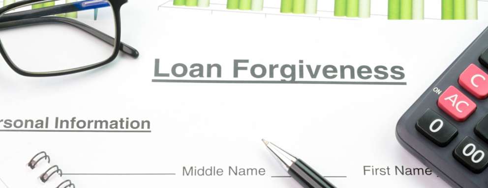 Concept of loan forgiveness. A calculator, a pencil, and a pair of glasses resting on top of a piece of paper that says "loan forgiveness" on it.