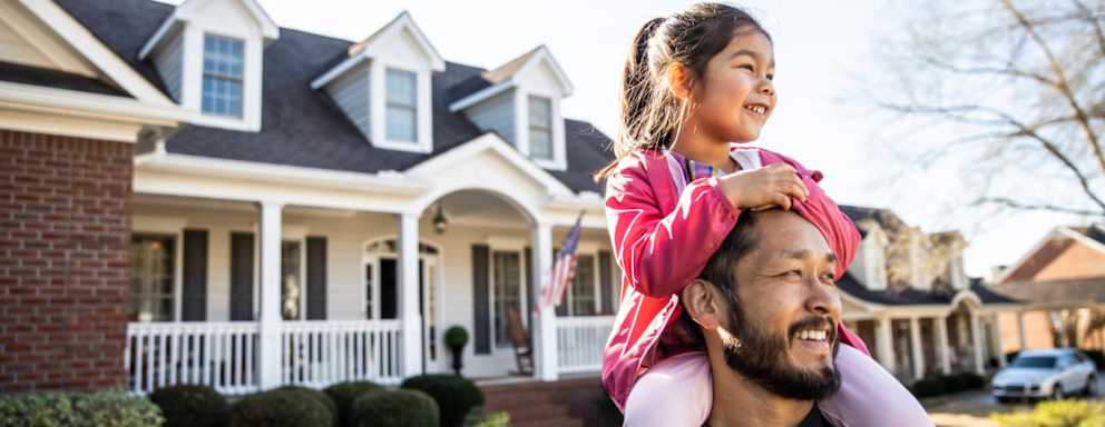 A smiling Asian American father with his daughter on his shoulders stands in front of his newly purchased home in a suburban neighborhood.
