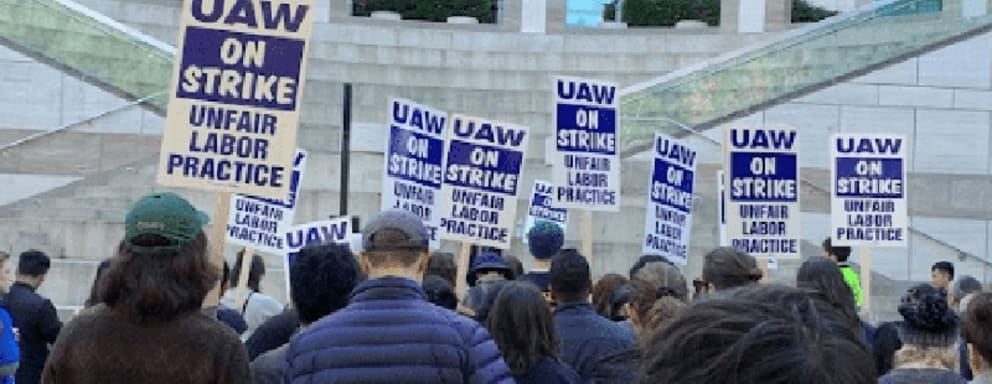 A group of protesters standing out in front of a building with signs that read "UAW on strike, Unfair Labor practices".