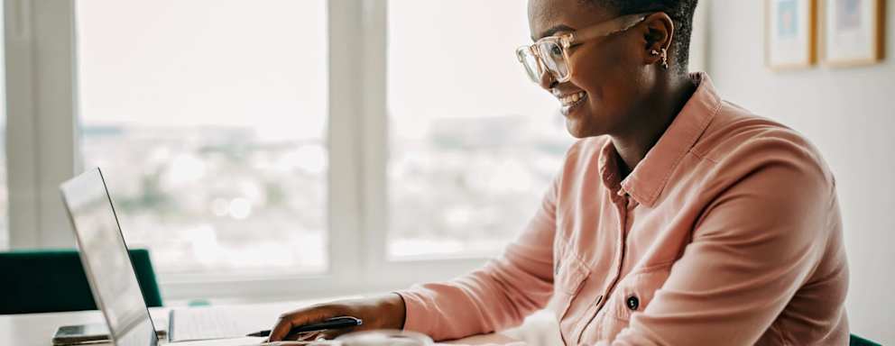 A woman working from home on her laptop. She is sitting at her desk by a big bright window.