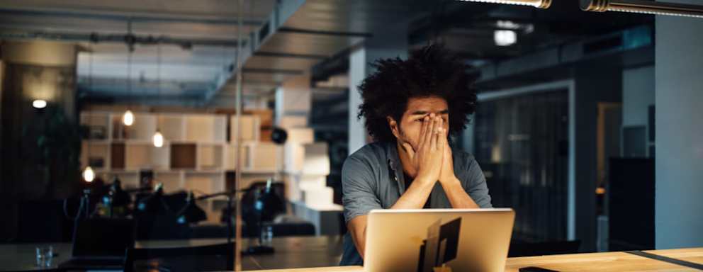 A young Black male employee sitting at his desk in a corporate office late at night. He has his hands on his face and his laptop is open.