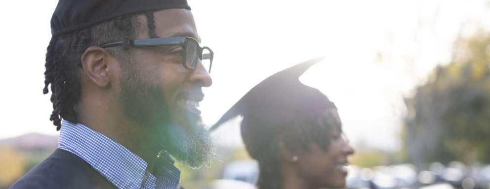 Adult persons who have just graduated university. They are both wearing cap and gowns and smiling while facing forward listening to the speaker.