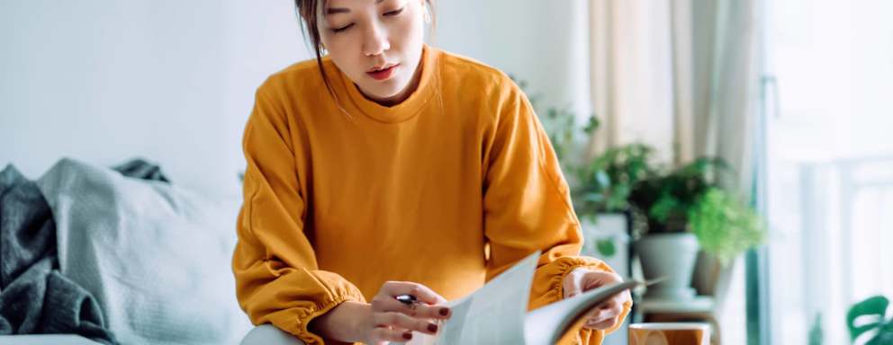 Female Asian American college student sitting at home on her living room couch. She is concentrating on studying for the LSAT exam. She is reading from an exam practice book and taking notes.