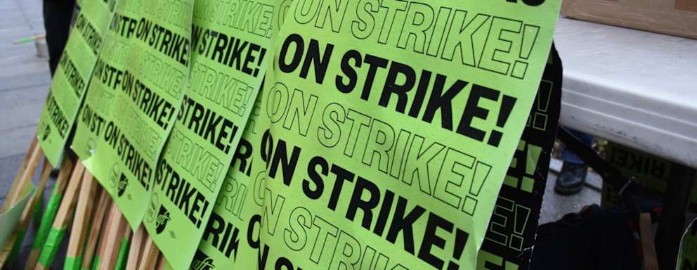Printed student strike signs resting against a sidewalk bench on the Temple University campus. The signs read, "TAs and RAs On Strike!"