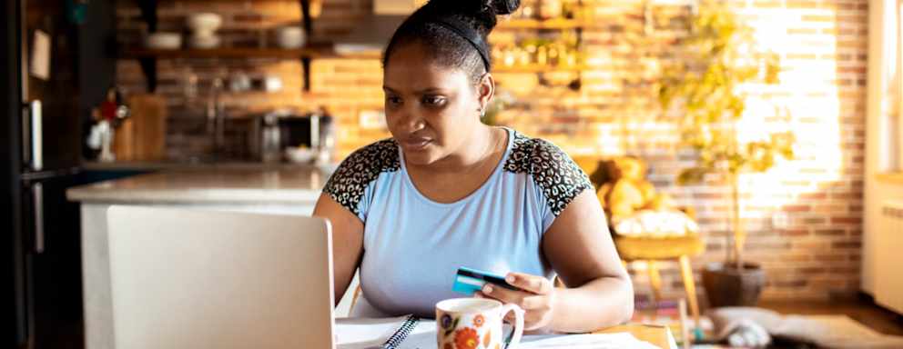 Female college student sitting at her dining room table in her apartment. She is using her credit card to make a tuition payment on her laptop computer.