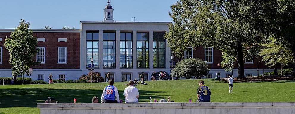 A view from Charles street of the grassy "Beach" and Milton S Eisenhower Library of the Johns Hopkins University; three male students, their backs facing the camera, sit on top of the marble university sign, other students sunbathing and playing catch on the grassy hill; the clock tower of Gilman Hall, a humanities building, peeks above the top of the library; Baltimore, Maryland, March, 2014. Courtesy Eric Chen. (Photo by JHU Sheridan Libraries/Gado/Getty Images).