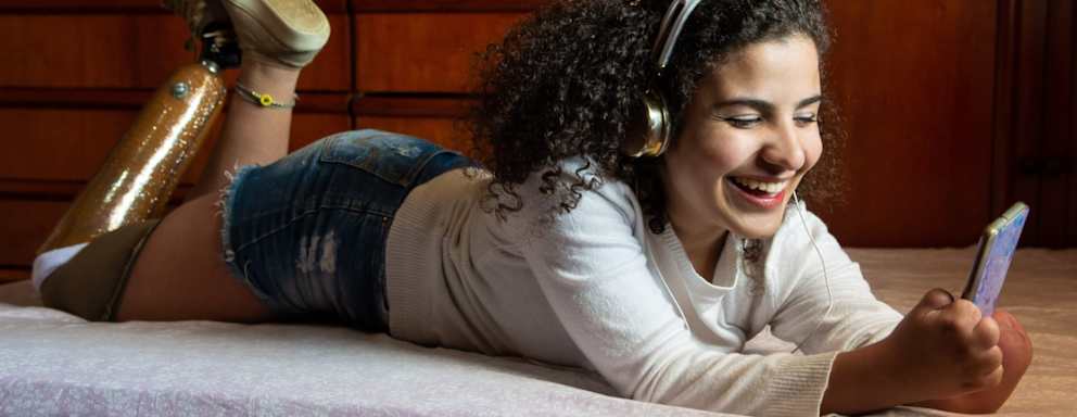 Female college student with prosthetic leg and symbrachydactyly hanging out on her bed in her dorm room. She's smiling while listening to music on her cellphone.