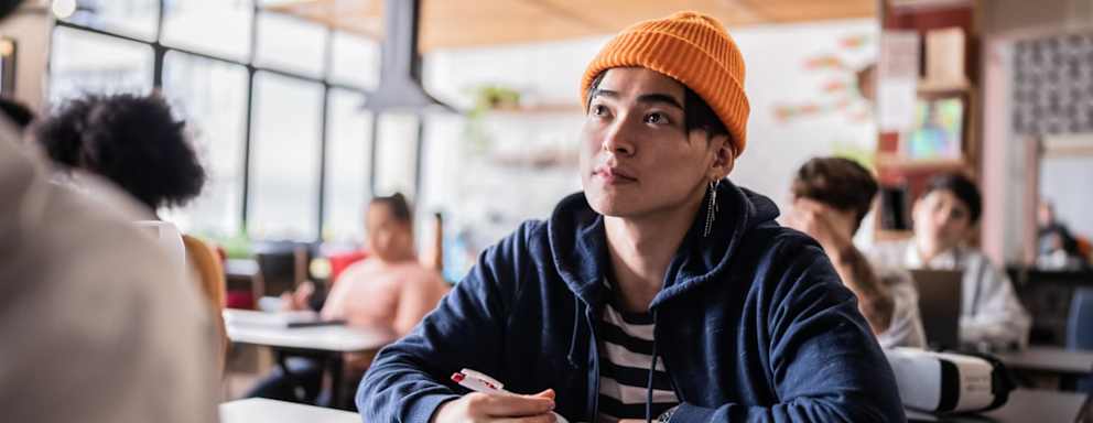 Asian American male college sitting at a table in a university lecture room. He is looking off camera to his professor speaking, while writing notes during class.