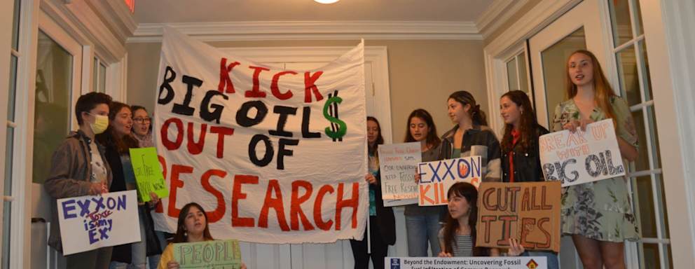 A group of college students standing in a living room holding protest signs related to getting rid of big oil.