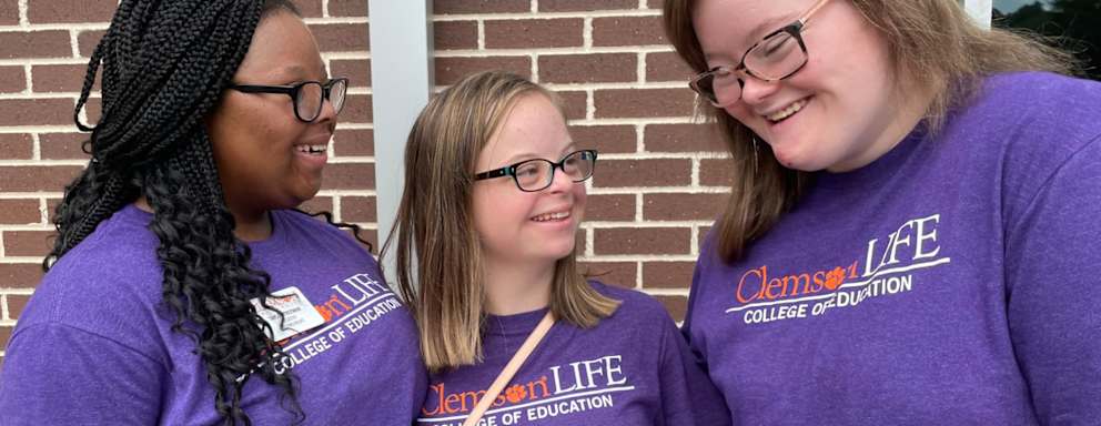 A photo of students standing outside smiling and talking with each other. They are all wearing "Clemson Life College Education" purple T-shirts.