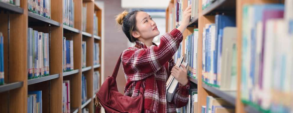 Female Asian American college student looking for textbooks on the shelves in her university's library.