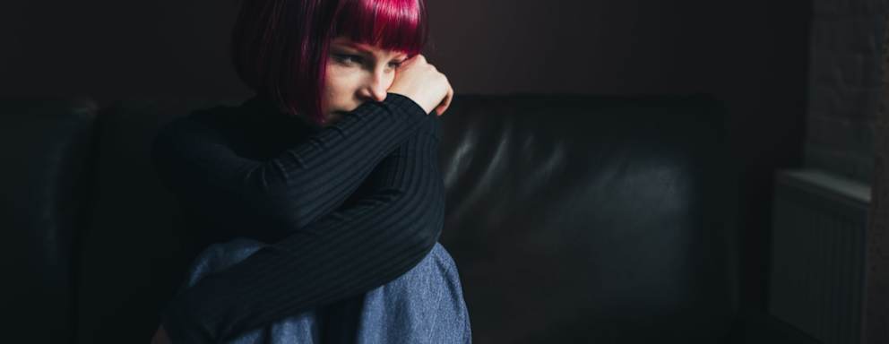 Sad female college student sitting alone on her living room couch with the lights off. She is hugging her knees to her chest and staring wistfully outside the window.