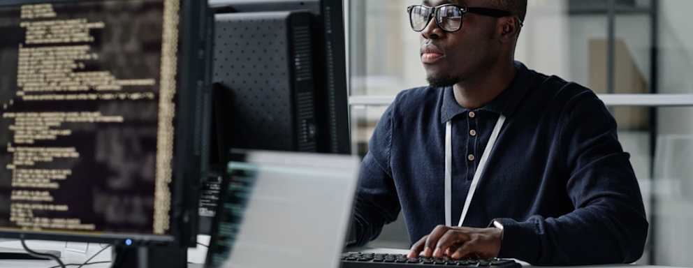 Black male cybersecurity engineer sitting at his desk in a corporate office. He is typing code for an internal security program on his desktop computer.