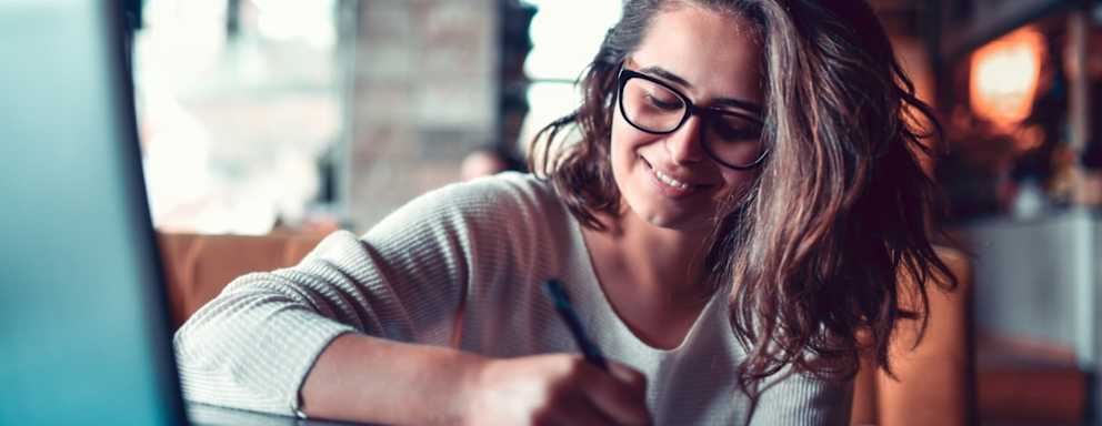 Female Taking Notes For Work Project While Researching Topic On Laptop