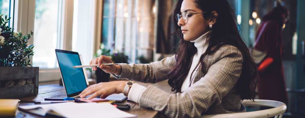 Female law student wearing glasses sitting at a table in a cafe. She is writing notes on a digital tablet. She is studying from several open textbooks.