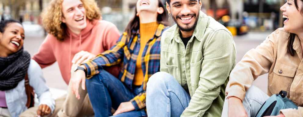 A multicultural group of male and female college students hanging out in a city park after class. They are sitting on a park bench, laughing over a shared joke with each other.