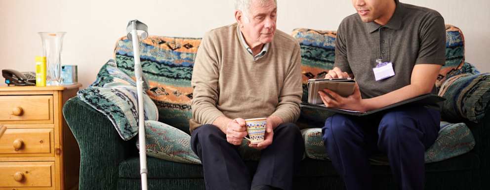 Black male social worker paying a house visit to a senior male client. They are sitting on the client's couch in his living room and chatting together.