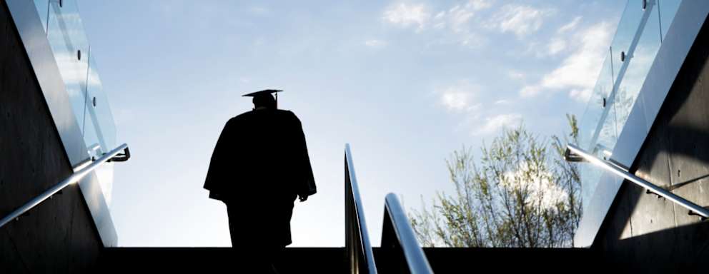 Silhouette of a college graduate in a cap and gown climbing up a set of steps on a university campus.