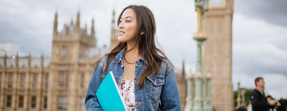 Asian American female college student standing on the Westminster Bridge in London, UK. She is holding a notebook. Big Ben can be seen in the background.