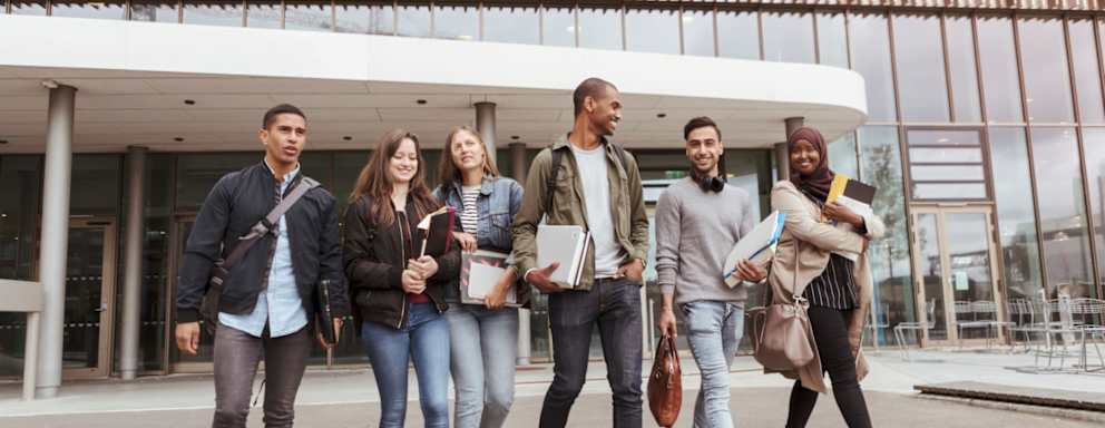 A diverse group of 6 college friends walking together on campus. They are walking out of a university building getting ready to hang out.