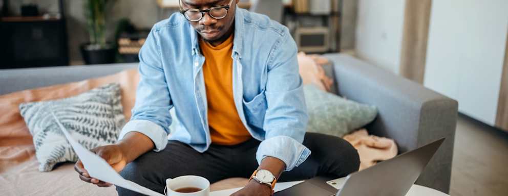Black male college student sitting on a couch in his living room. He has his laptop open on the coffee table, and is holding a paper bill in his right hand. He is typing numbers on a calculator to check the bill amount on his student loan bill.