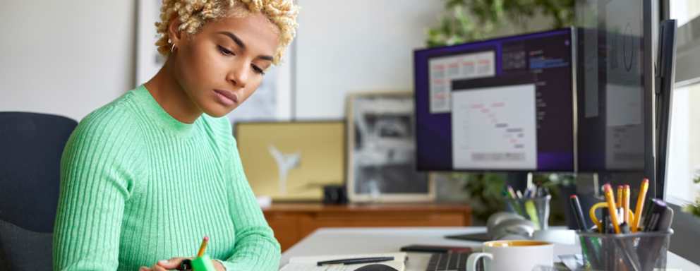 Black female business professional working from home at her desk. She is writing notes with a felt tip pen on paper while looking at charts and spreadsheets on her desktop computer.