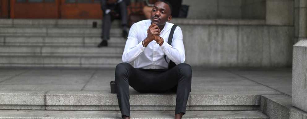 Black male lawyer sitting on steps. He is dressed in a business suit, and is looking offscreen with a thoughtful, serious look on his face.