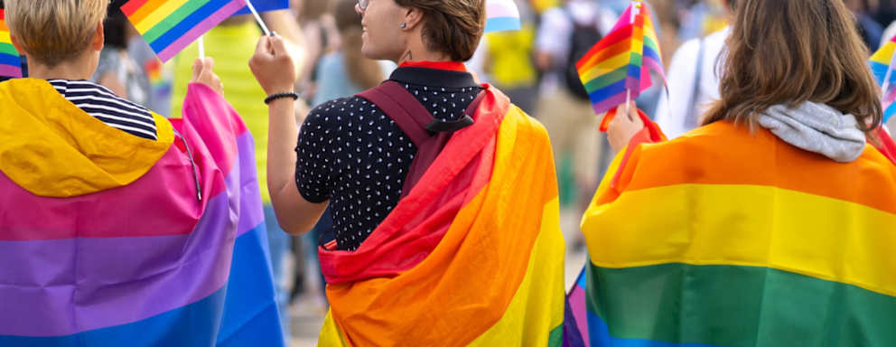 A group of three college students wrapped in bisexual and gay pride flags are waving small flags while watching a gay pride parade during Pride Month.