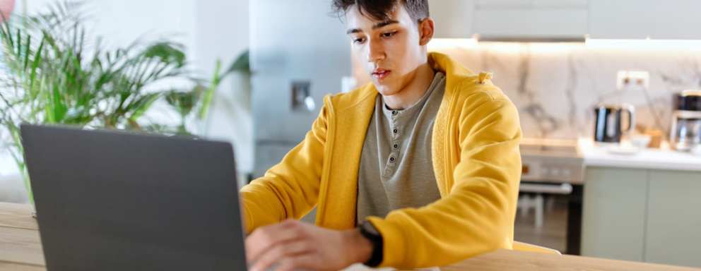 Male high school student sitting at a dining table at home. He is typing his college essay on his laptop computer.