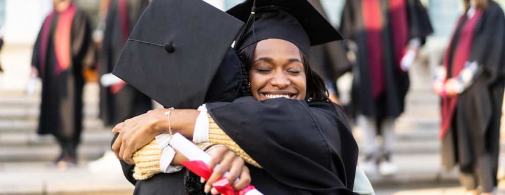 Two Black female college students are hug each other after receiving their diplomas on graduation day. They are wearing black caps and gowns and smiling.