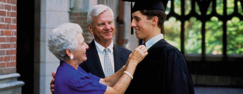 A Caucasian male college student dressed in a black cap and gown celebrates with his grandparents on his graduation day. His grandmother is adjusting his tie, while his grandfather gives him a congratulatory pat on the back.