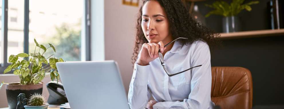 Female accountant sitting at a desk in her office. She has taken her glasses off, and is reading documents on her laptop computer.