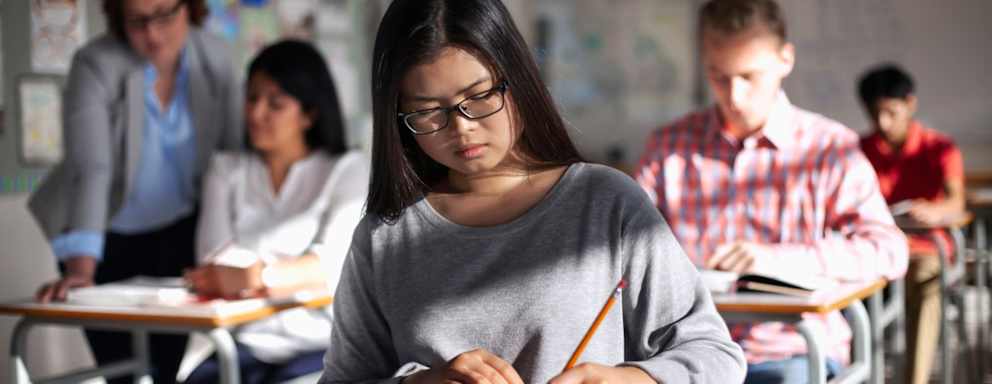 Asian American female student taking a standardized text in a high school classroom. She is sitting at her own desk, surrounded by several other students taking the exam.