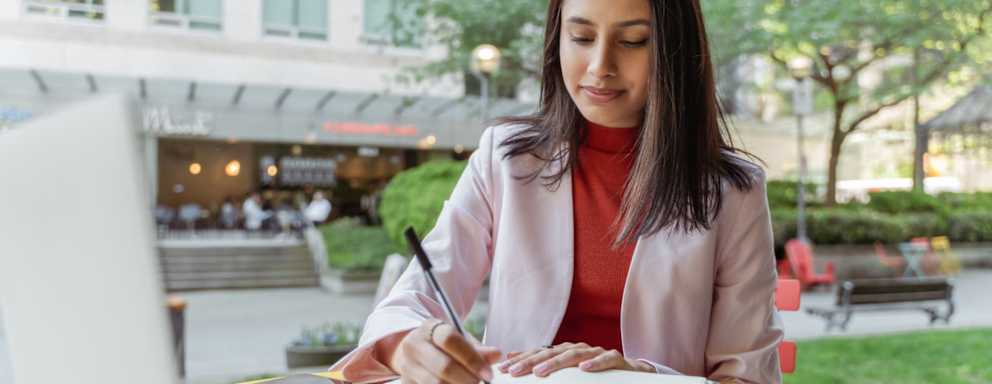 Female South Asian business student sitting at a table in an outdoor park. She is writing notes while referencing study materials on her laptop computer.