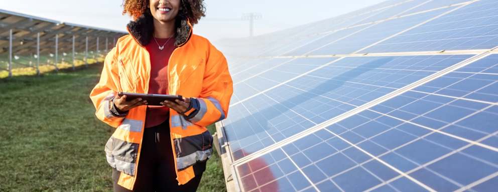 Black female technician in reflective clothing holding a tablet device is standing by a solar grid at a solar farm and smiling.