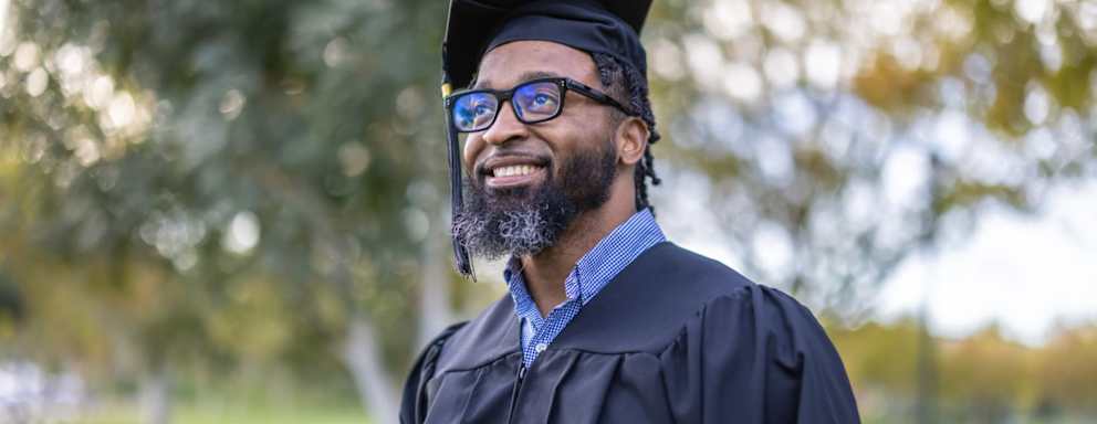 Happy Black adult male college student wearing a black cap and gown at his commencement ceremony.
