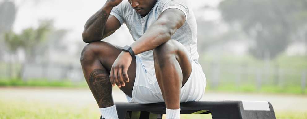 Black male college football player sitting on the field. He is rubbing the temples on his forehead, and has an exhausted look on his face.