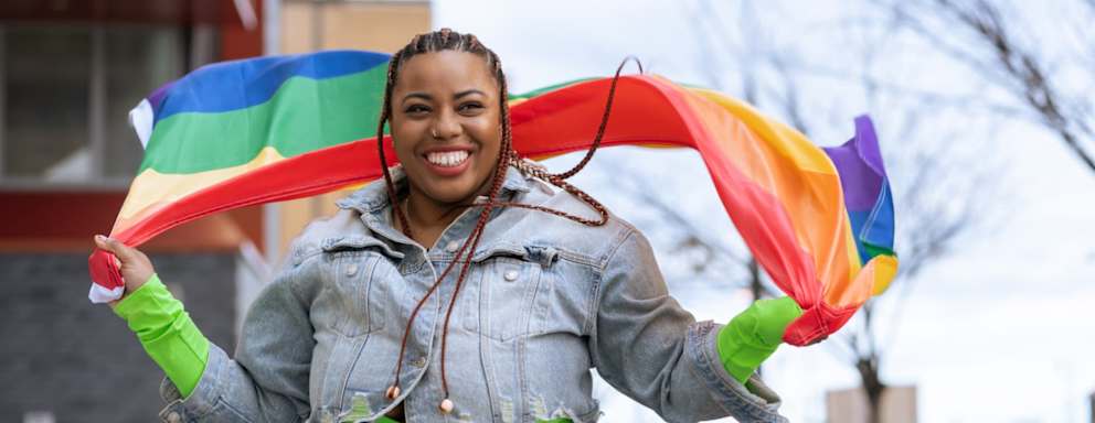 Black female college student sitting outdoors holding a rainbow flag during an LGBTQIA+ pride celebration.