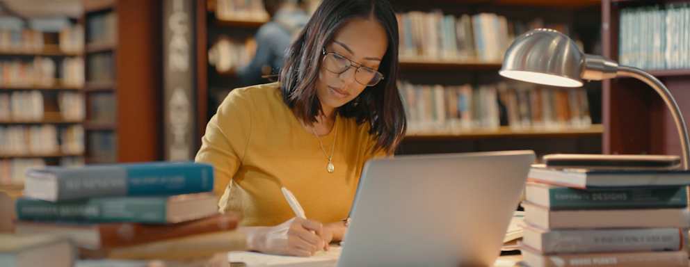 Female graduate student sitting at a table in a university library. She has her laptop open and is writing notes in her notebook to study from later.