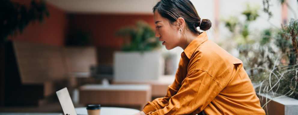 Female Asian American businesswoman working on her laptop in a modern co-working space, surrounded by green plants.