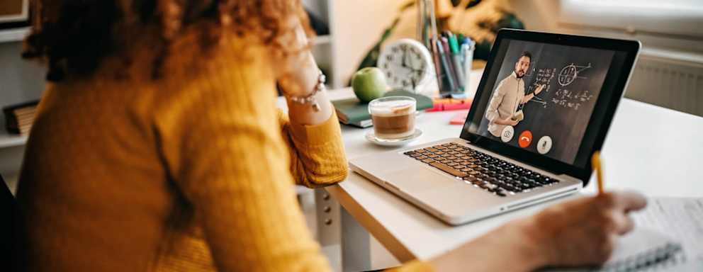 Caucasian woman sitting at a desk in her apartment. Her laptop is open, and she is taking notes on a notepad. She is taking a class online: her professor appears in a Zoom meeting on her laptop, and is demonstrating math concepts on a blackboard behind him.